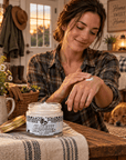 A woman in a plaid shirt applies cream from a jar labeled 'Whipped Cloudy Pastures Beef Tallow ' on her hand.  Made by Amish Country Soap Company